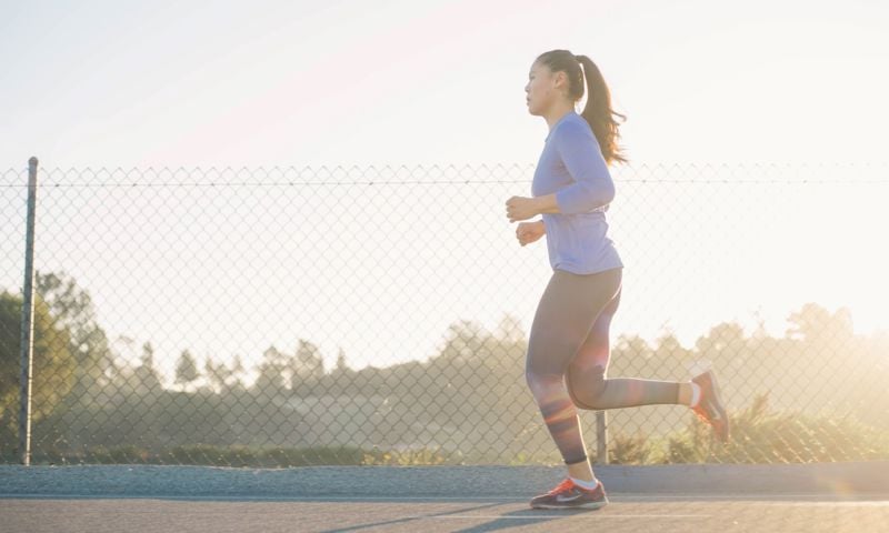 A woman in activewear jogging along a road in the sunlight.