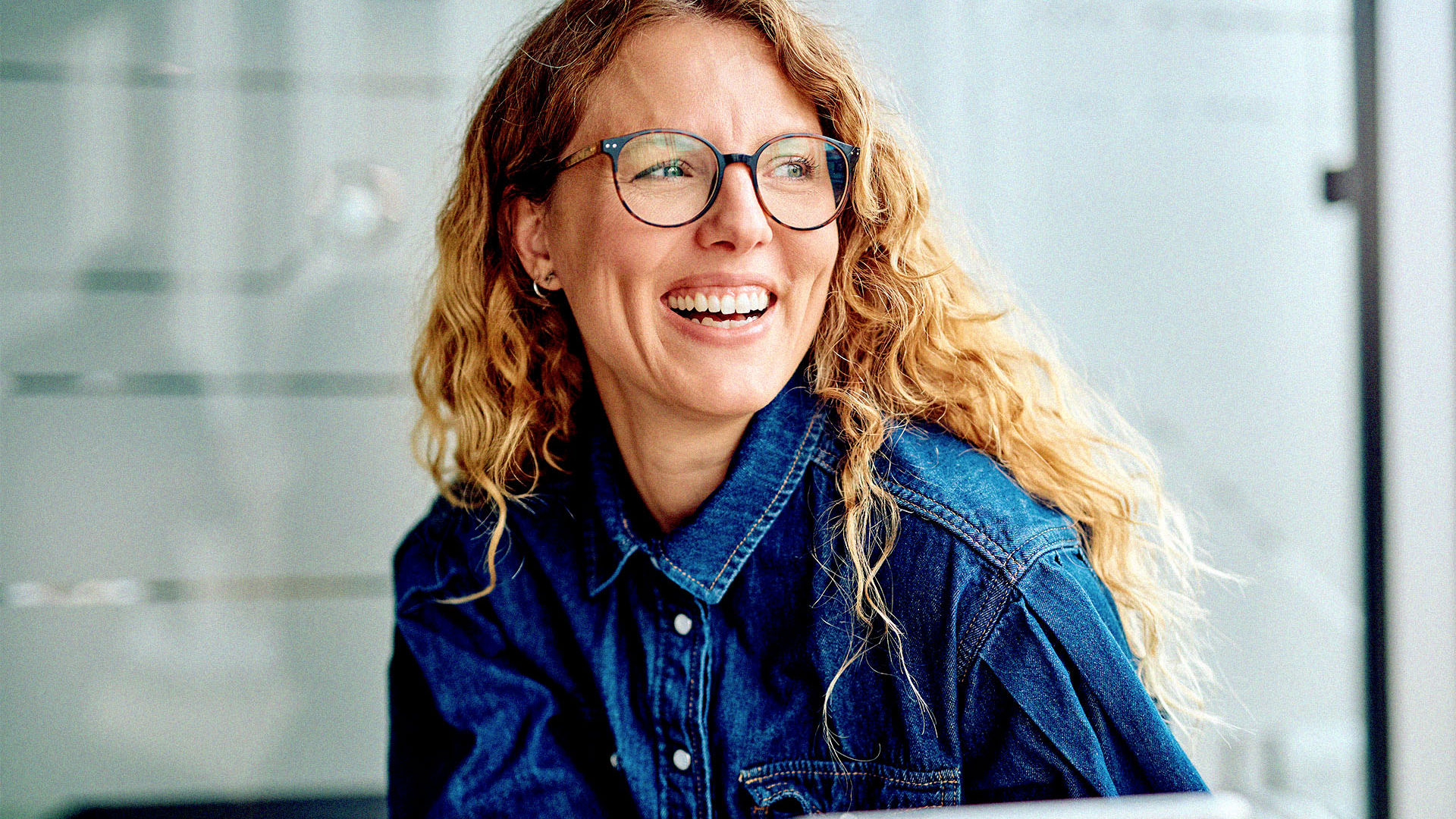 Young lady wearing denim and stylish glasses smiling across a table.