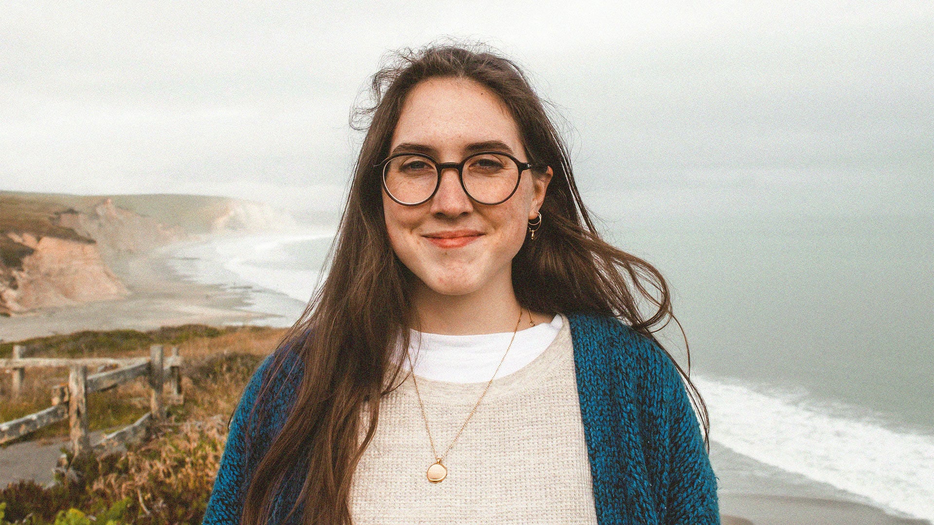 Young female with glasses smiling standing on clifftop with ocean in background